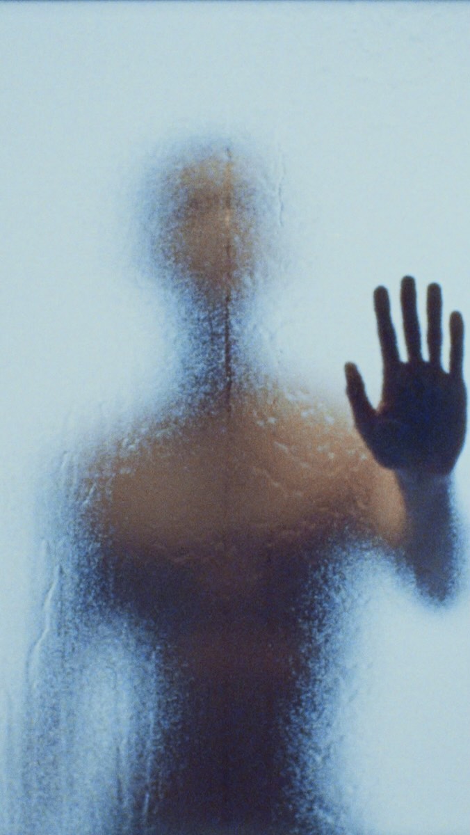 Abstract, backlit silhouette of an adult figure against a steamy glass surface with visible handprint in a blue-toned, textured scene.