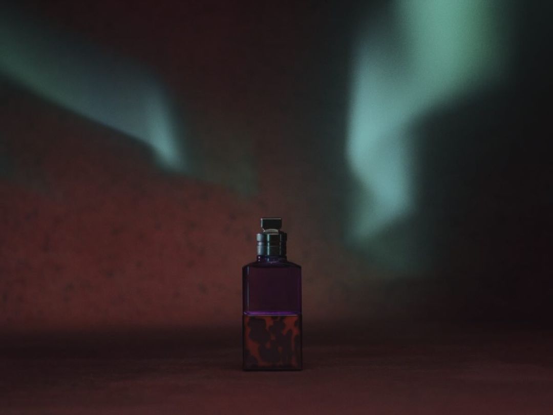 Elegant still life of a square purple glass bottle filled with dark liquid, set against a blurred backdrop of soft light patterns.