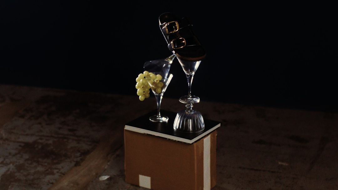 Still life featuring a black sandal balanced atop two martini glasses and grapes, presented on a box against a dark background.
