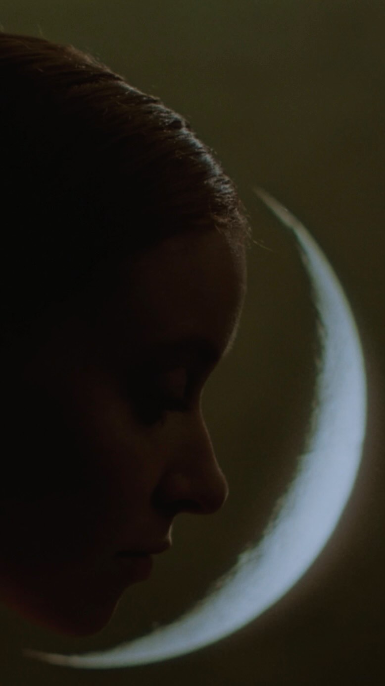 Dramatic portrait of an adult woman's face in deep shadow against a glowing crescent moon background in a vertical format image.