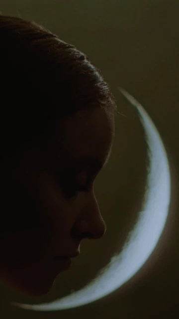 Dramatic portrait of an adult woman's face in deep shadow against a glowing crescent moon background in a vertical format image.