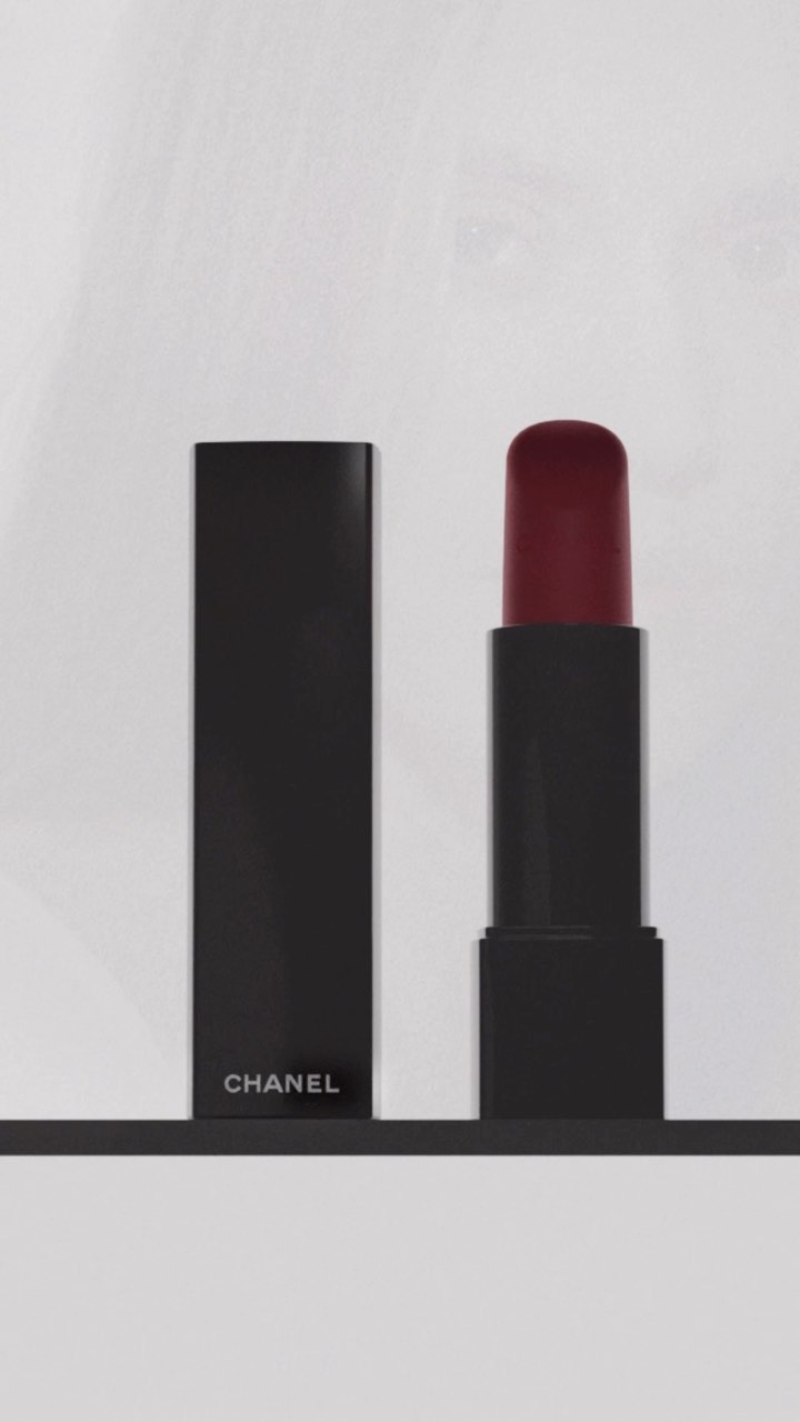 A black lipstick tube with opened cap and a lipstick bullet with burgundy color are seen against a white background and dark shelf.