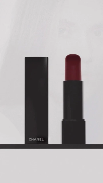 A black lipstick tube with opened cap and a lipstick bullet with burgundy color are seen against a white background and dark shelf.