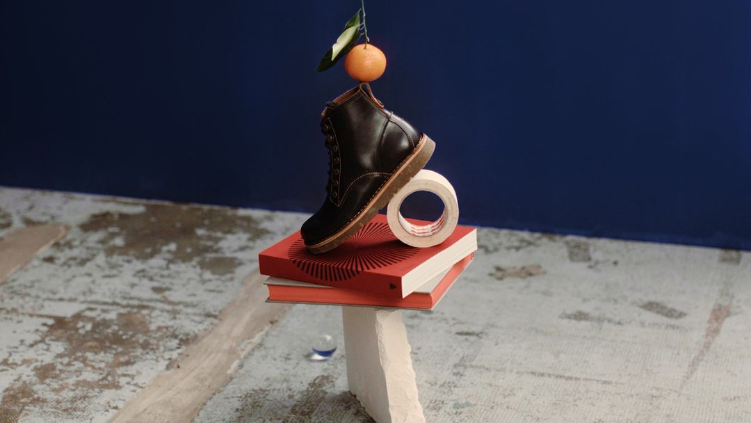 Still life composition with a leather boot balanced on books, tape, and a white stone, topped with a fresh orange against a blue background.