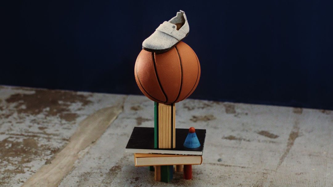 Artistic still life shot of a brown basketball topped with a grey shoe, balanced on books and a badminton birdie on a concrete surface.