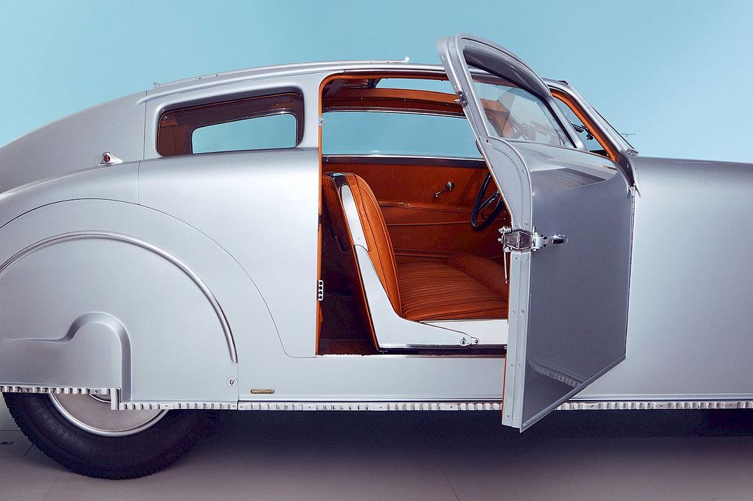 Silver vintage car with an open door revealing the interior. The car is photographed in a studio against a solid light blue background.