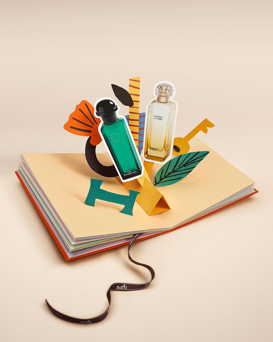 Green and gold perfume bottles emerge from an opened book in a studio setting; paper cut-out art is beside the bottles.