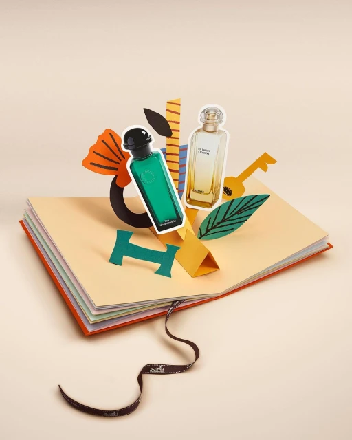 Green and gold perfume bottles emerge from an opened book in a studio setting; paper cut-out art is beside the bottles.