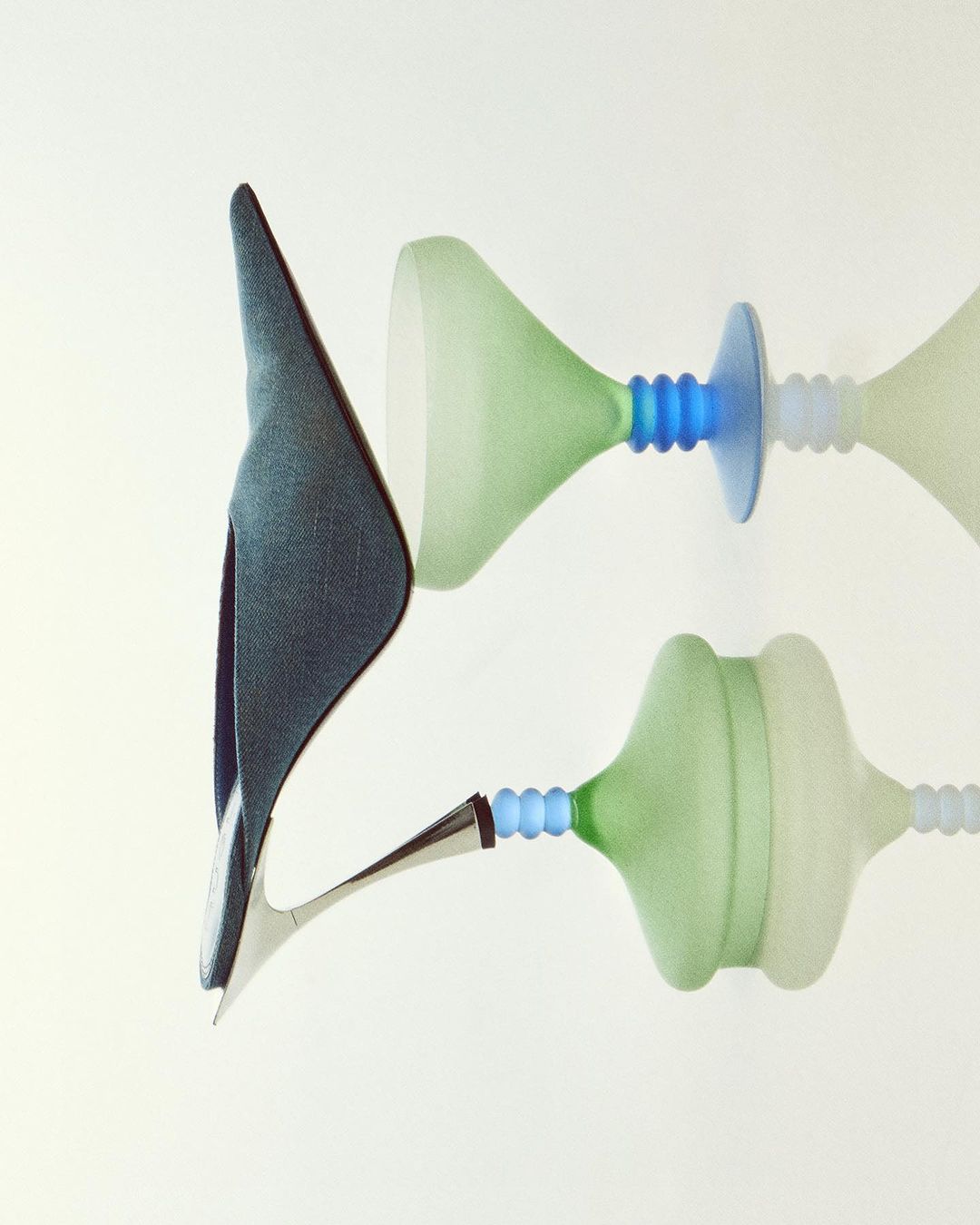Fashion still life showcasing a high heel shoe alongside abstract hourglass-shaped design objects in pale green, blue, and white against a white backdrop.