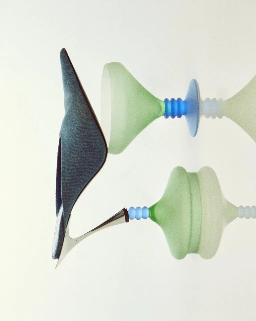 Fashion still life showcasing a high heel shoe alongside abstract hourglass-shaped design objects in pale green, blue, and white against a white backdrop.