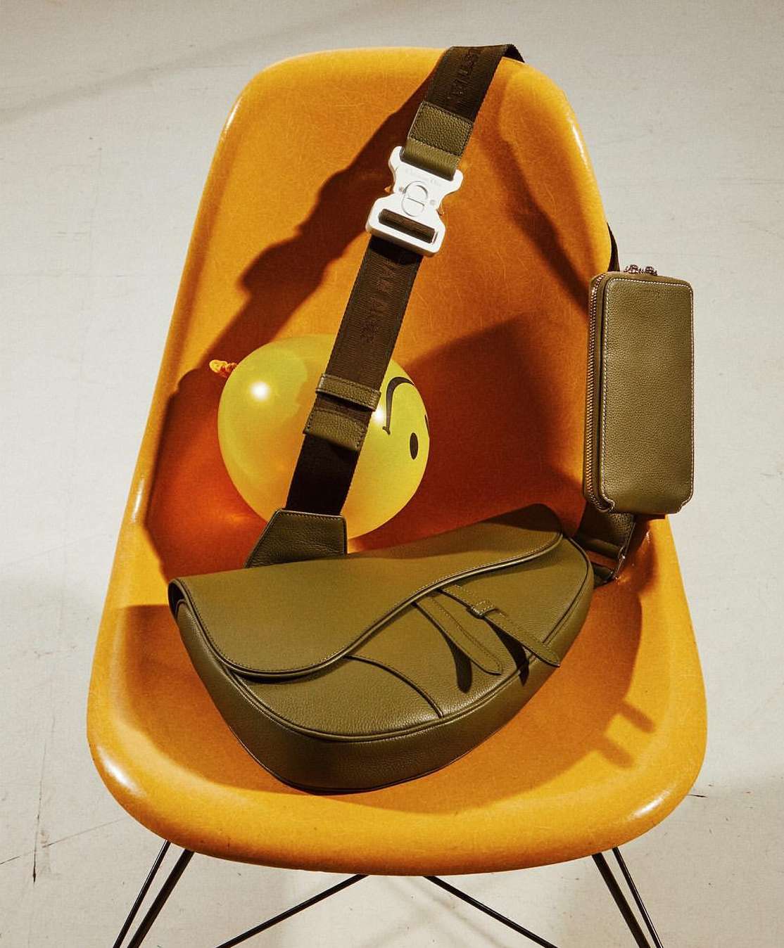 Saddle bag with strap over mustard-yellow molded plastic chair. Bag has an olive-green pouch and yellow balloon with a smiley face in the background.
