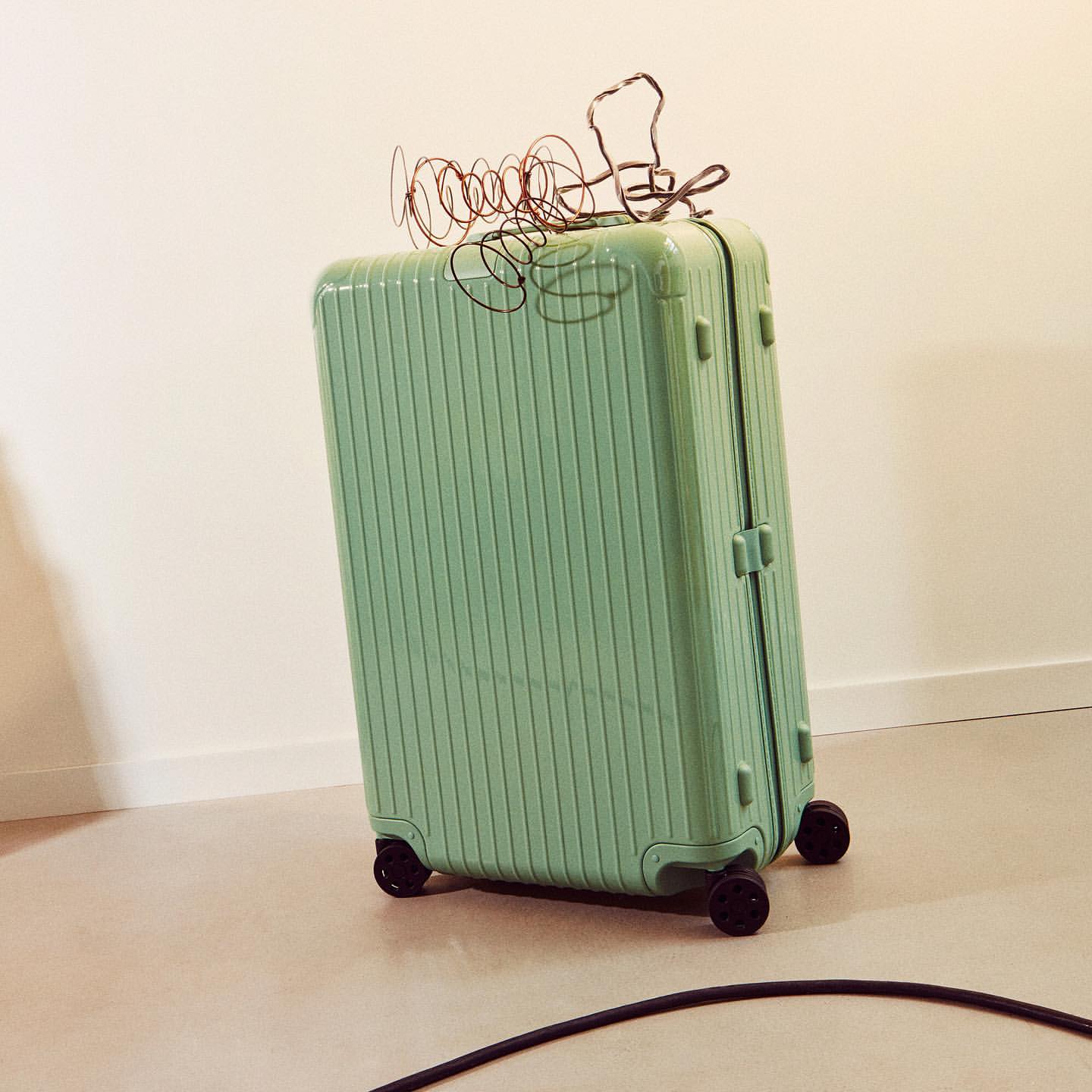 Mint green hardshell suitcase with black wheels and a tangle of brown wire on top sits against a neutral wall and floor.