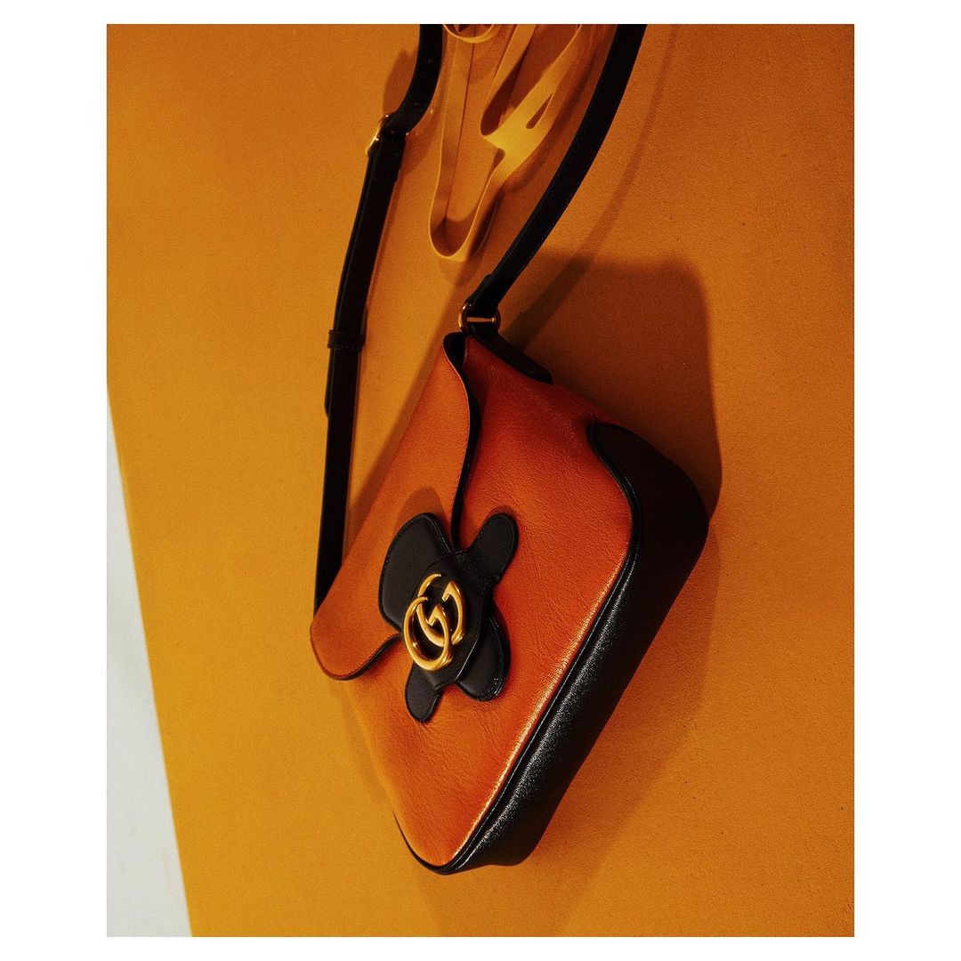Studio shot of an orange leather Gucci handbag with black flower detail, suspended against a matching orange backdrop. Fashion product.