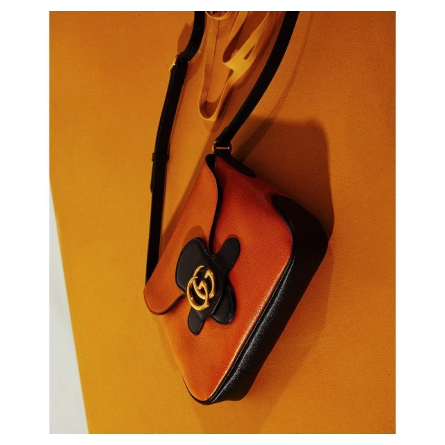 Studio shot of an orange leather Gucci handbag with black flower detail, suspended against a matching orange backdrop. Fashion product.