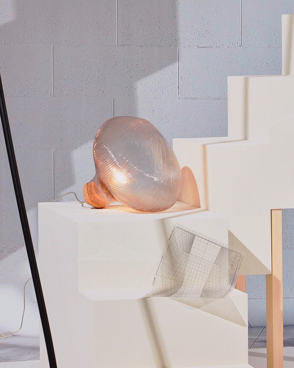 Illuminated wire mesh sculpture casting a warm glow on a white platform against a textured grey background.