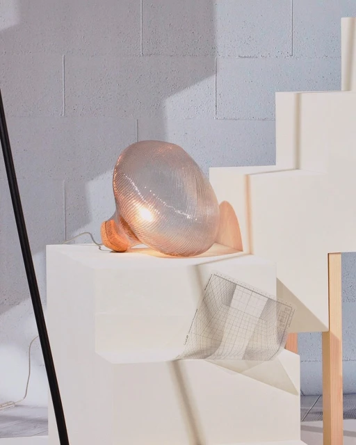 Illuminated wire mesh sculpture casting a warm glow on a white platform against a textured grey background.