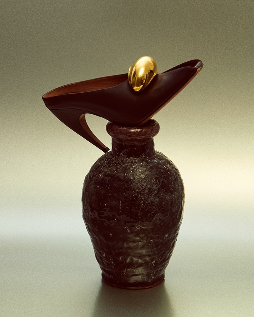 Still life of a black high heel shoe with a gold accent balancing on a dark textured ceramic vase, studio lighting, and light background.
