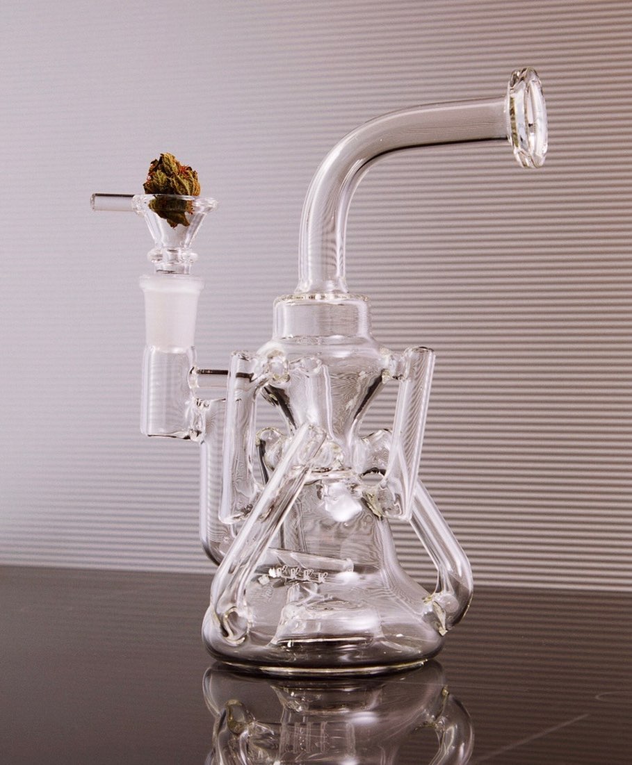 Clear glass bubbler on a black reflective surface set against a striped background, showcasing modern smoking accessory design.