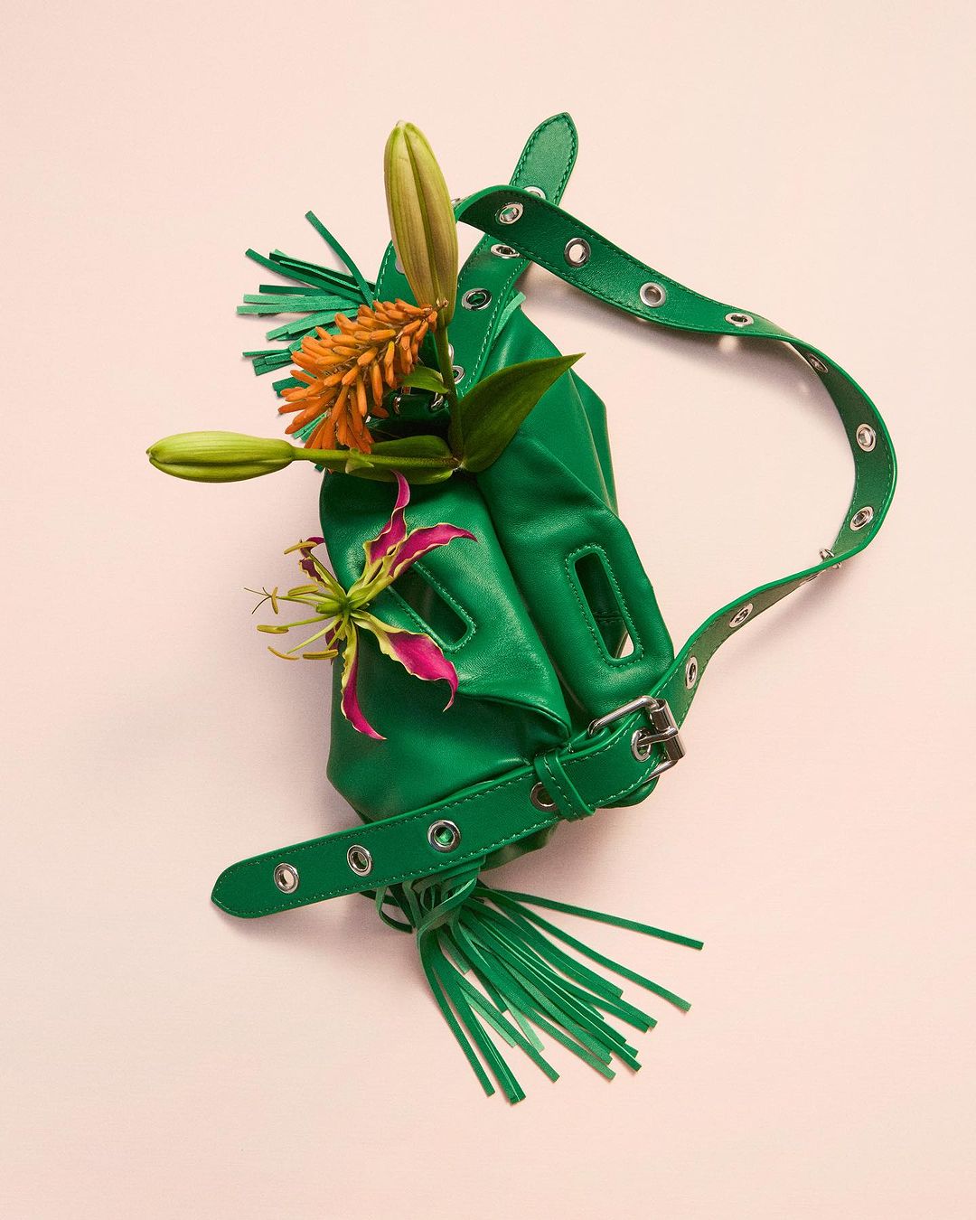 Fashionable green purse adorned with vibrant lilies, accented by a belted strap, and set against a soft pink backdrop in a studio setting.