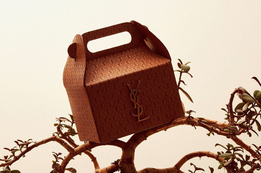 Brown textured gift box with gold logo emblem on a tree branch with a light beige background, angled view.