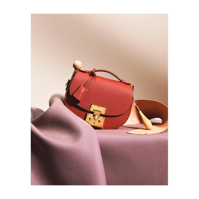 Red leather handbag with gold clasp, displayed on a mauve surface with matching draped leather and a light beige background.