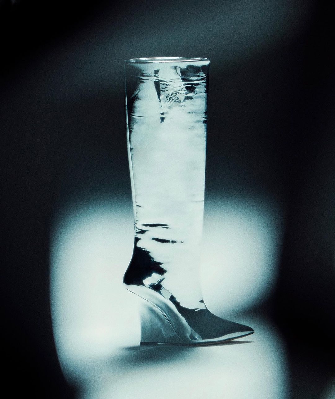 Modern glass boot with a wedge heel, displayed against a blurred, cool-toned background in a studio setting with professional lighting.