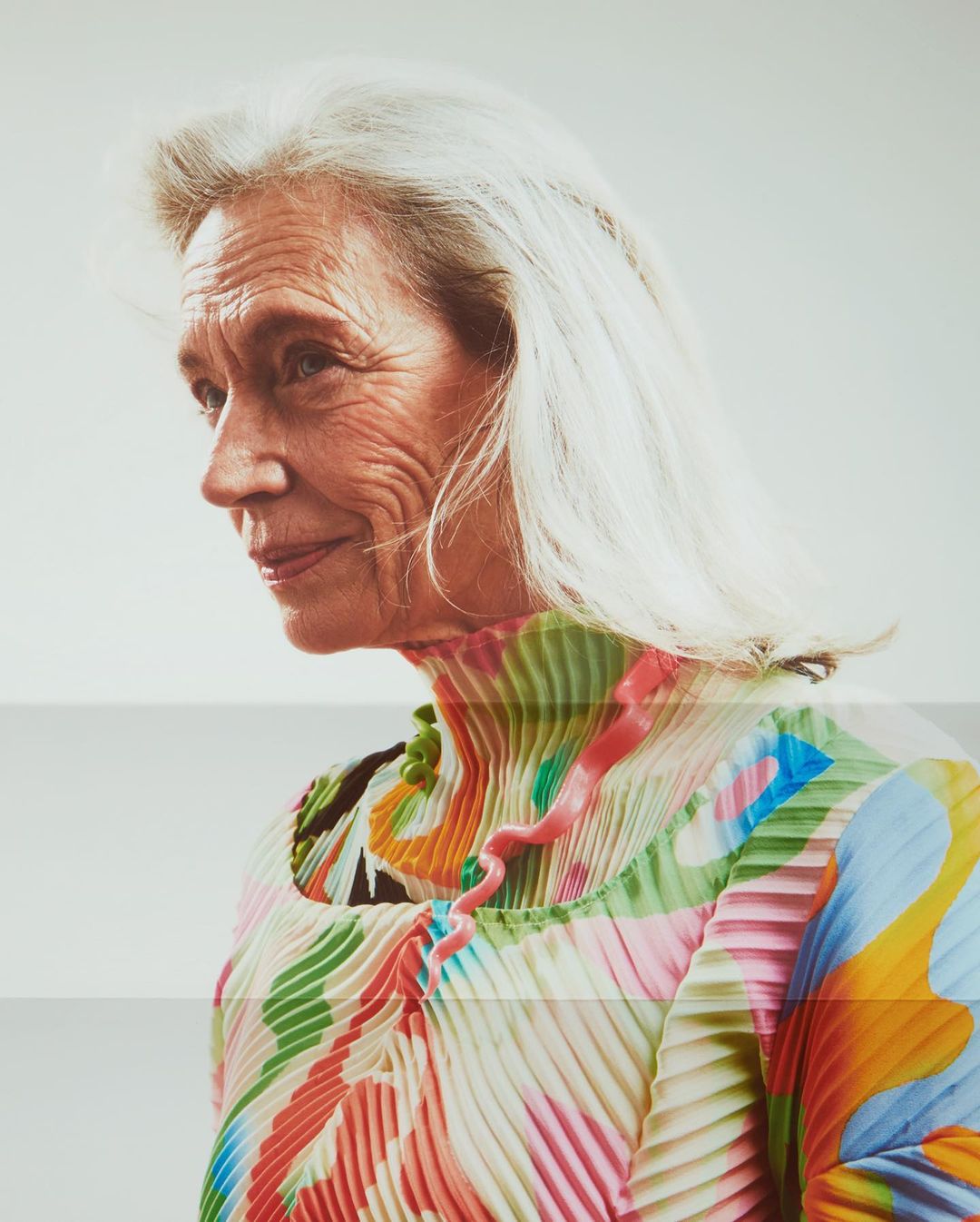 Elegant older woman in pleated dress. Portrait of a senior female with white hair wearing patterned top in light, airy studio setting.