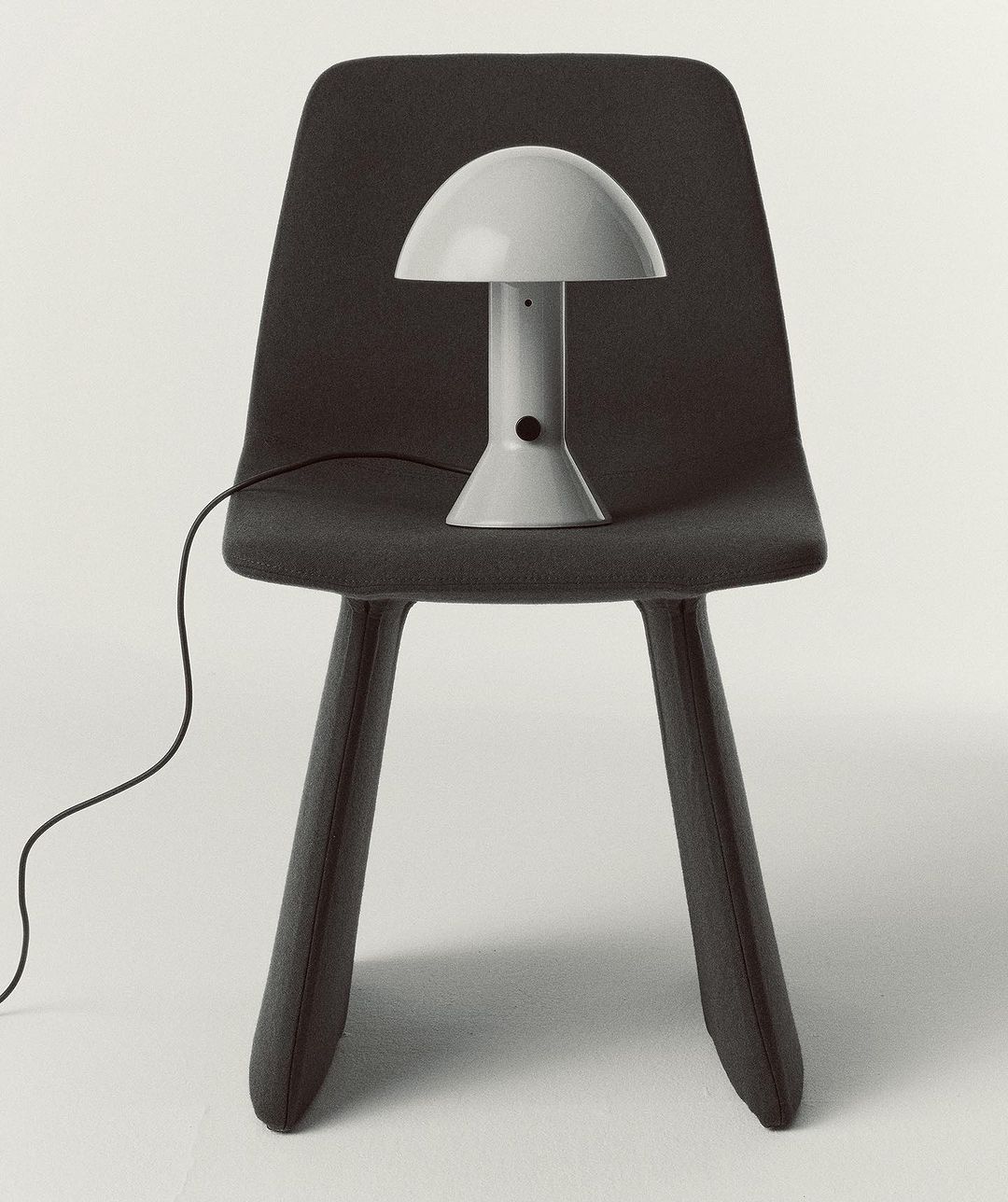 Grey mushroom lamp on a grey chair against a white background in an interior setting. The lamp's electrical cord is visible.