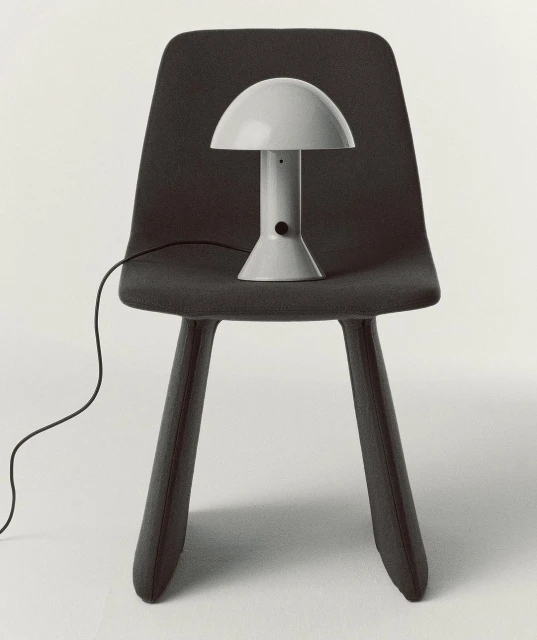 Grey mushroom lamp on a grey chair against a white background in an interior setting. The lamp's electrical cord is visible.