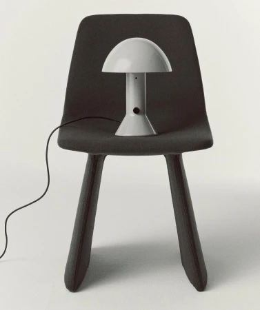 Grey mushroom lamp on a grey chair against a white background in an interior setting. The lamp's electrical cord is visible.