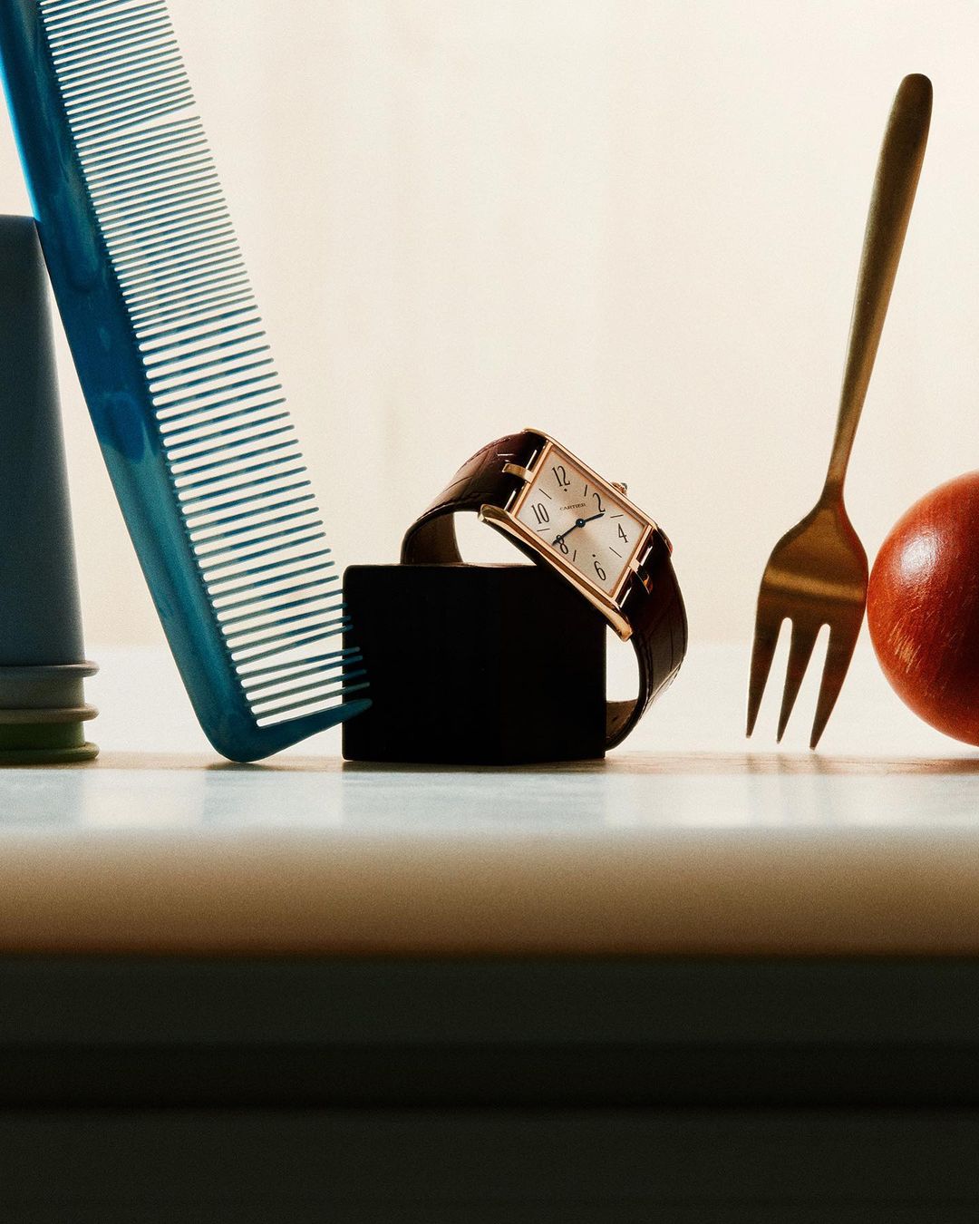 Still life photography featuring a wristwatch, comb, fork, and apple on a white surface, set against a neutral background.