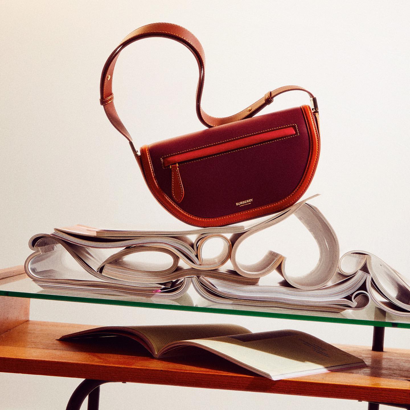 Burgundy leather Burberry shoulder bag with tan trim and strap, presented on a glass table with curated magazines and subtle studio lighting.