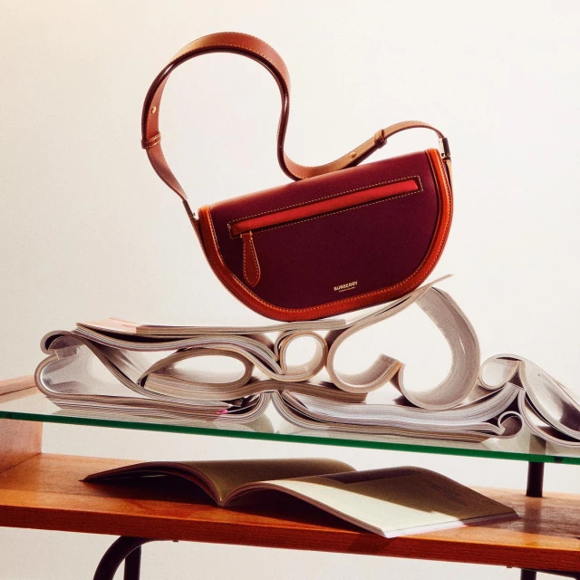 Burgundy leather Burberry shoulder bag with tan trim and strap, presented on a glass table with curated magazines and subtle studio lighting.