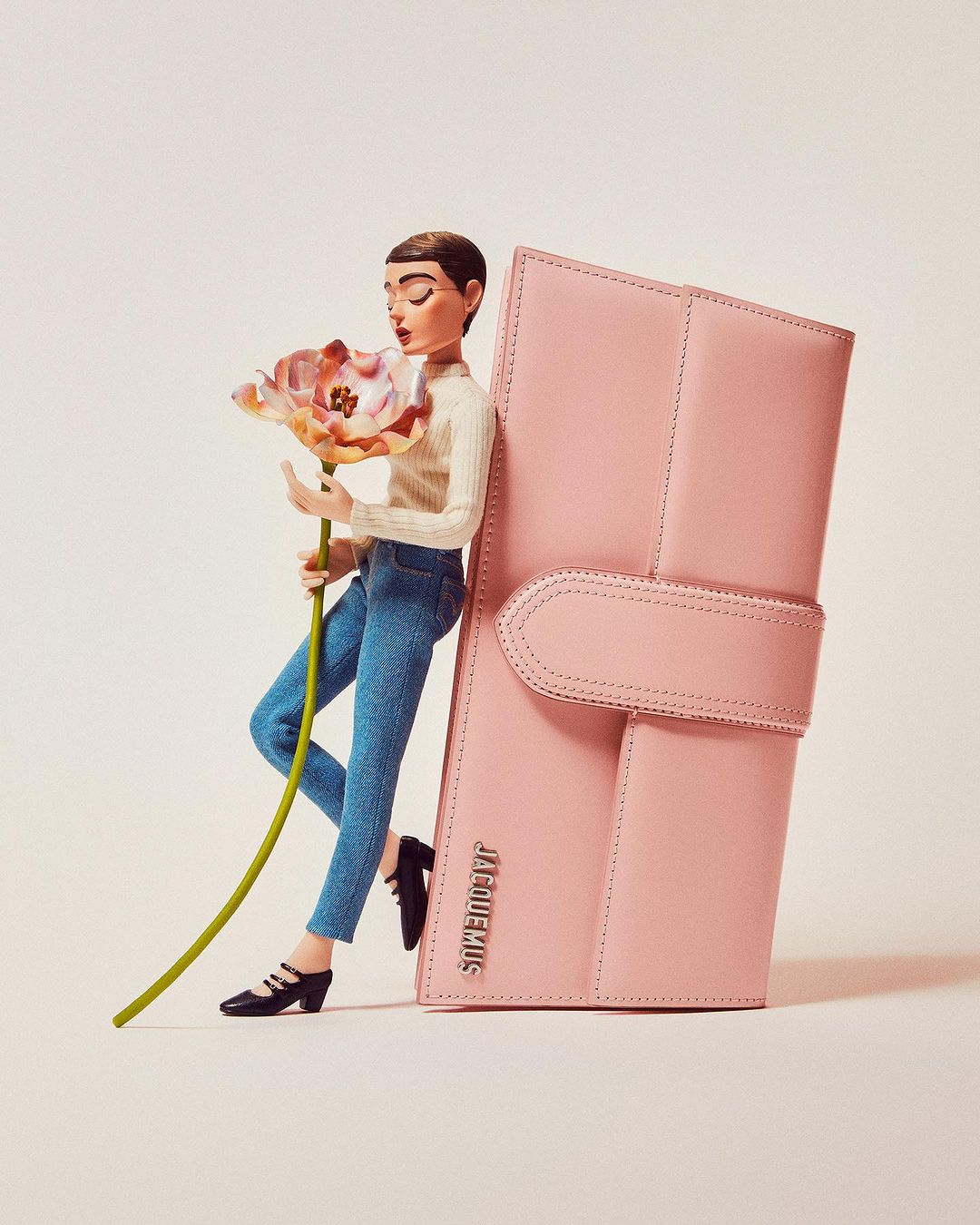 Conceptual fashion shot featuring a posing figure with a large pink leather handbag and a large flower on a light pink studio backdrop.
