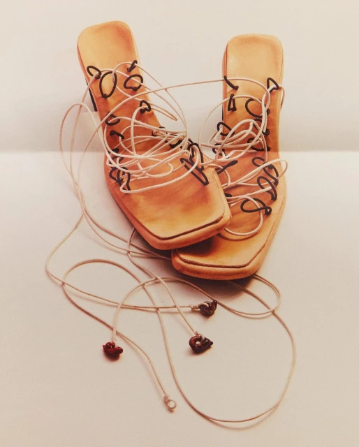 Pair of tan leather high-heeled sandals with long string laces on a white surface, conveying a fashion-forward and minimalist aesthetic.