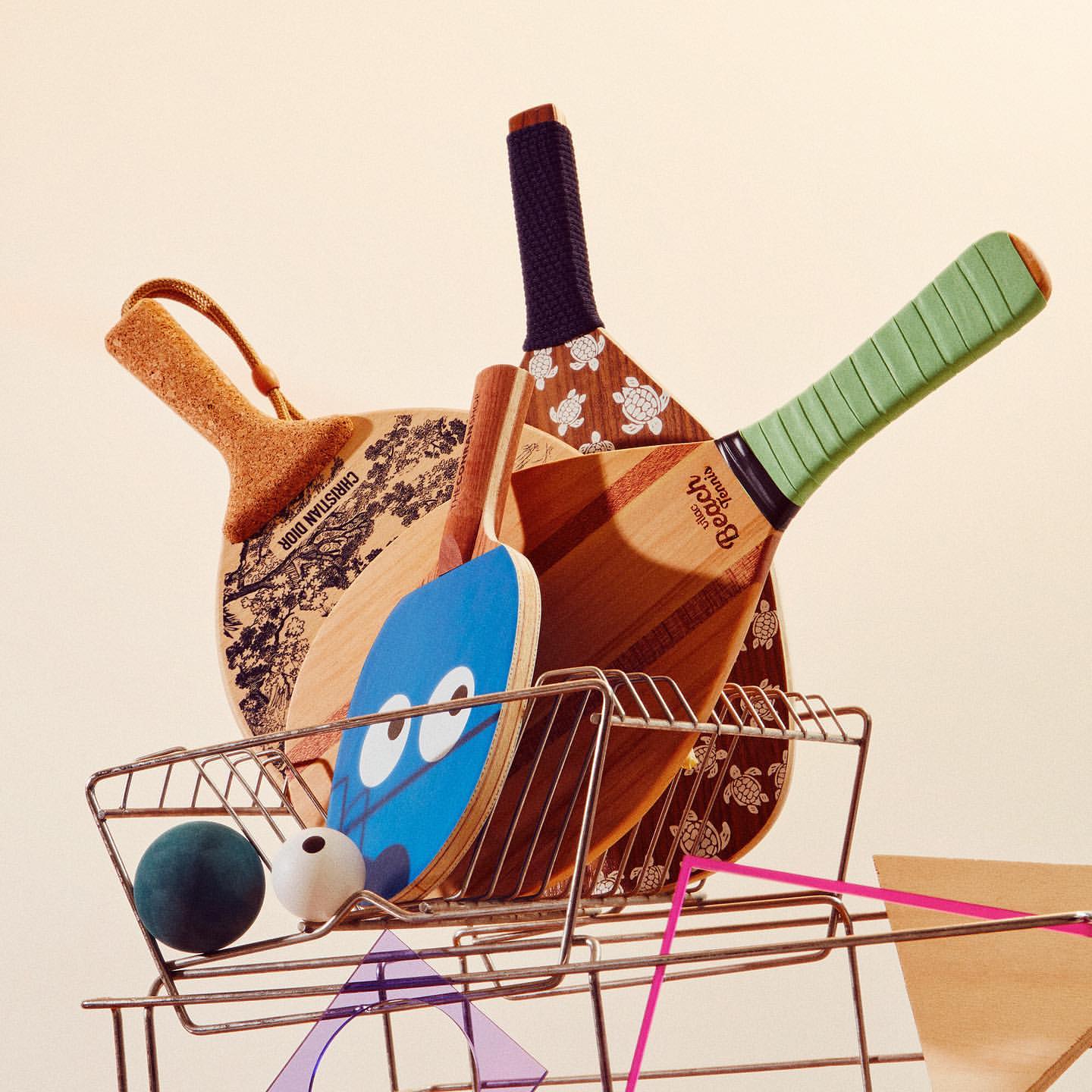 Arrangement of wooden beach paddles with varied designs in a wire basket, accompanied by colorful balls and geometric paper shapes.