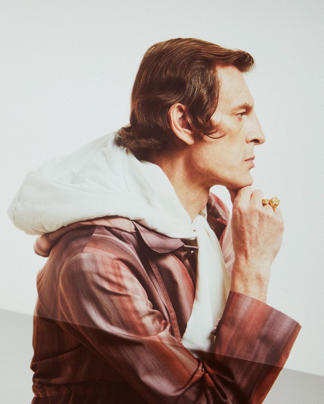 Side view of a man in a striped coat with a white hooded scarf, hand to his chin, featuring a gold ring against a blurred white background.