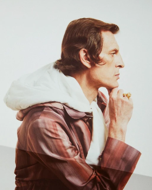 Side view of a man in a striped coat with a white hooded scarf, hand to his chin, featuring a gold ring against a blurred white background.