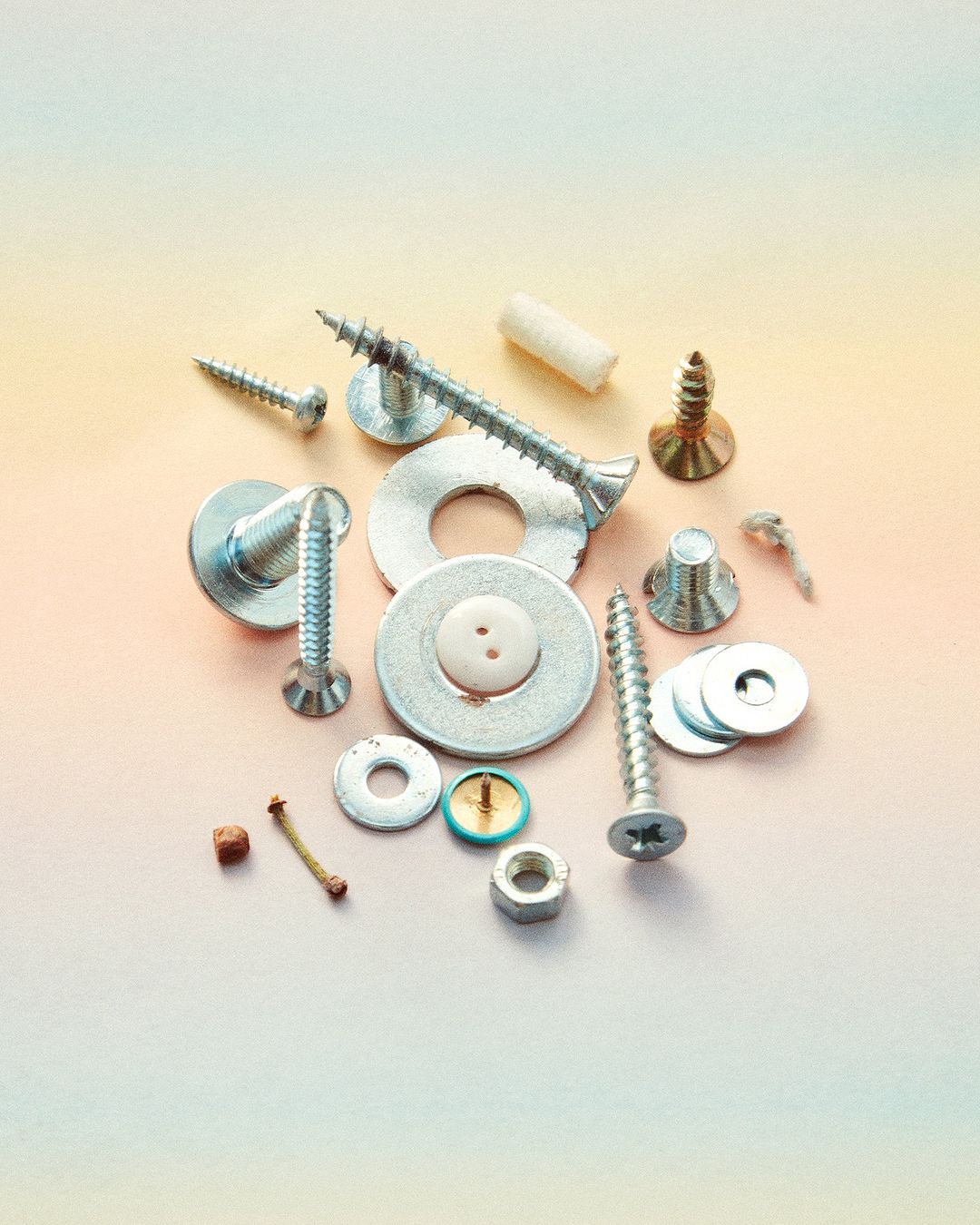 Close-up of various silver-toned metal screws, nuts, bolts, washers, and fasteners scattered on a gradient peach and light green surface.