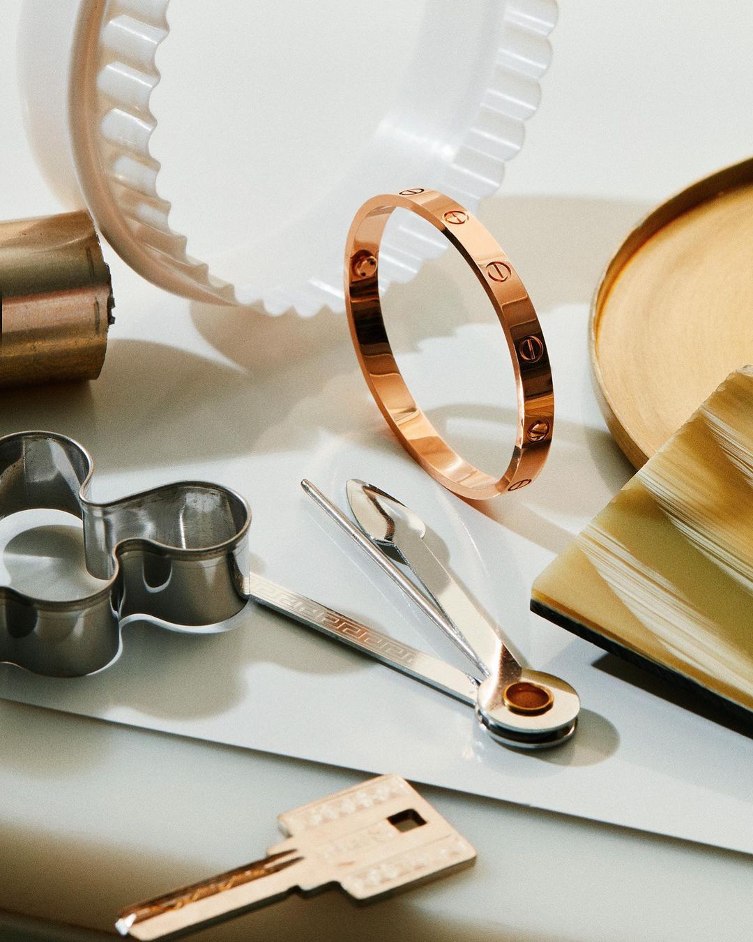 Still life with a Cartier bracelet, metal kitchen gadgets, a tray, and a key arranged on a white surface, product photography.