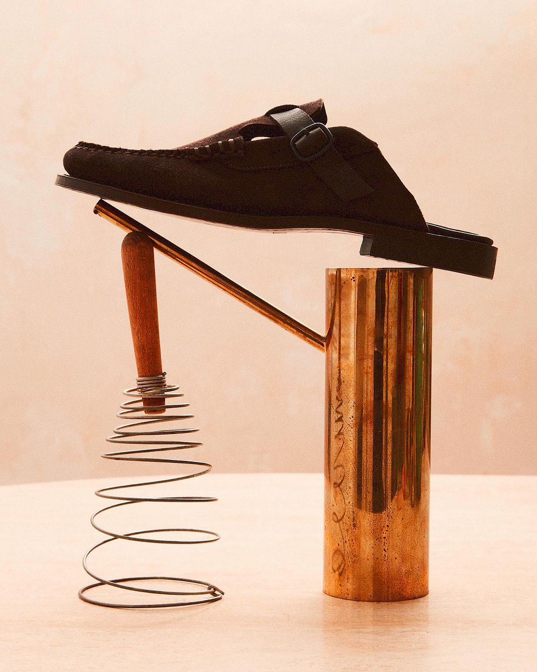 Studio shot of a brown, buckled, slip-on loafer resting on a copper watering can and a metal whisk against a peach-colored background.