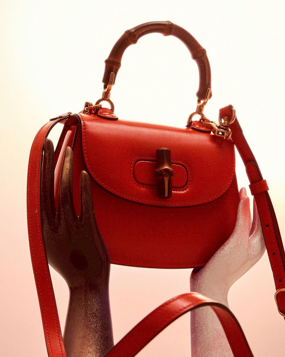 Red Gucci Diana handbag with bamboo handle on display with two metallic hands holding it up against a bright white background.