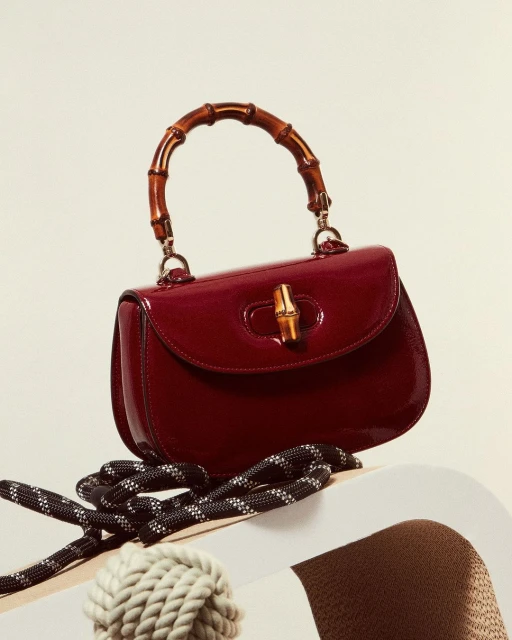 Burgundy patent leather Gucci Bamboo 1947 mini top handle bag with bamboo handle, striped rope strap, and beige background.
