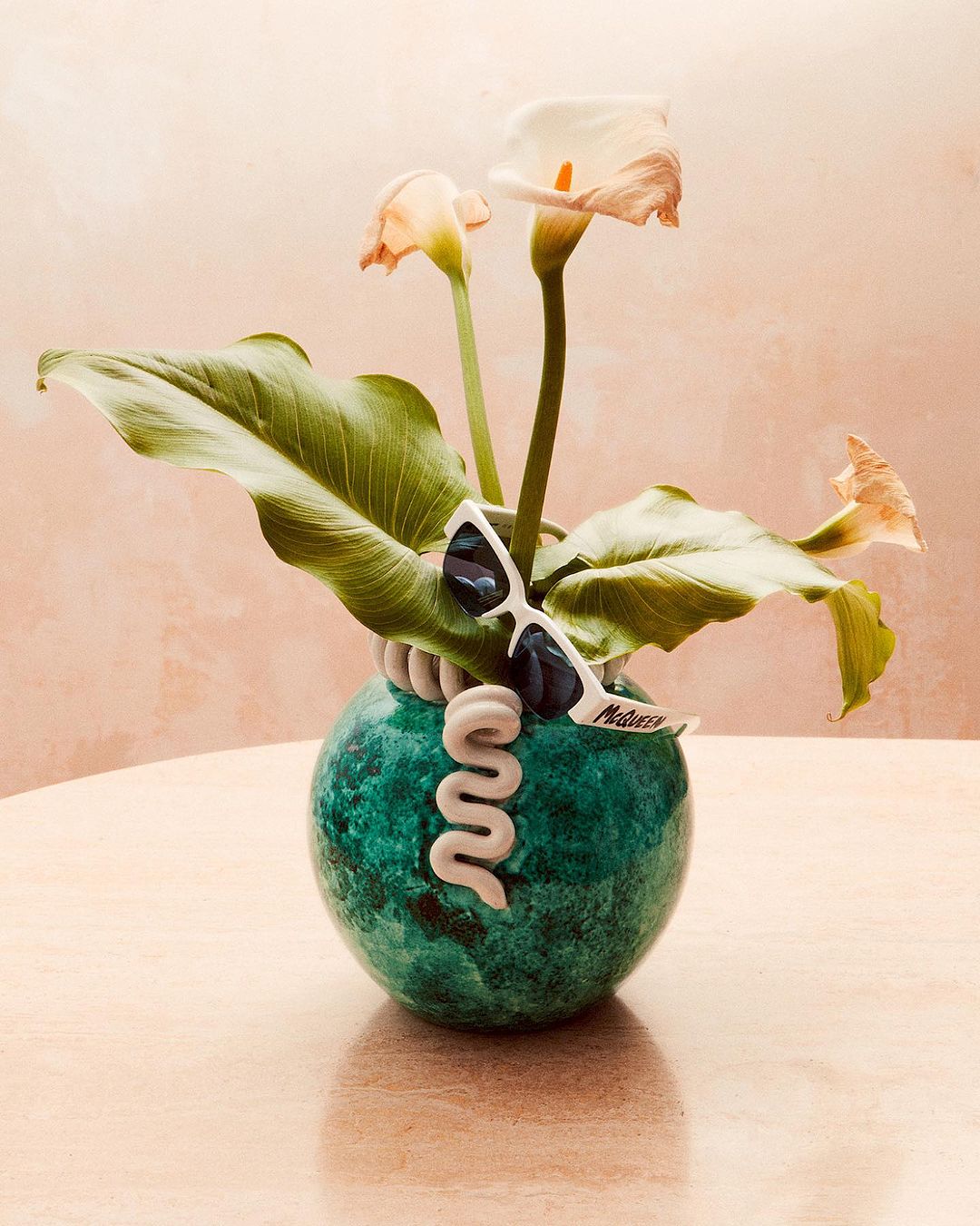 Still life of calla lilies in a decorative vase with sunglasses on a beige table against a peach-colored wall in a brightly lit room.