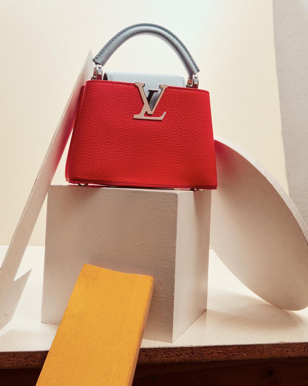 Red leather handbag with a silver logo, short grey handle, sitting on a white cube with geometric shapes against a neutral background.