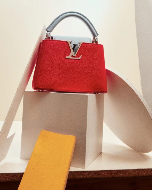 Red leather handbag with a silver logo, short grey handle, sitting on a white cube with geometric shapes against a neutral background.