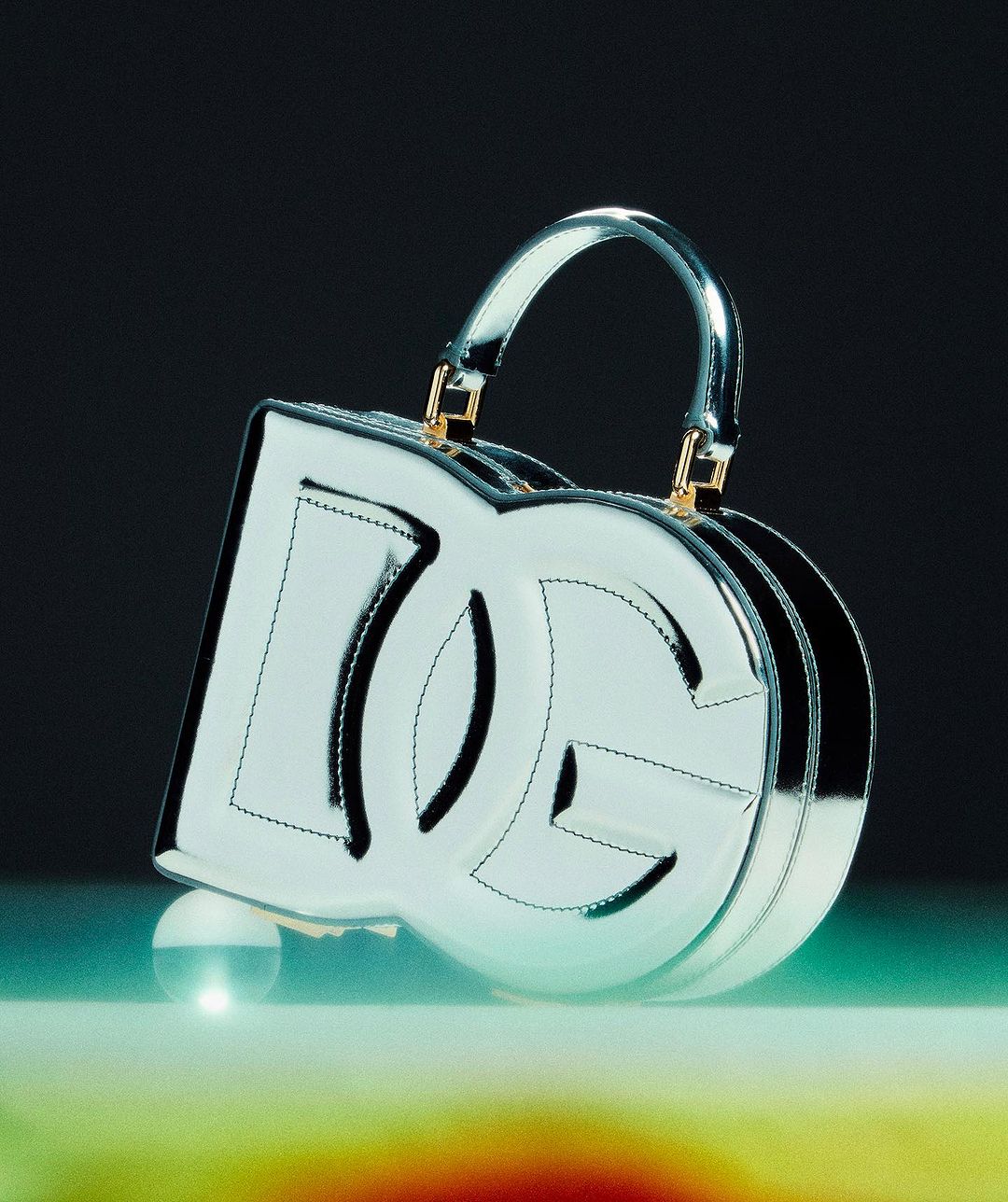 Dolce & Gabbana glossy silver handbag featuring logo cutout with top handle on a colorful reflective surface against dark background.