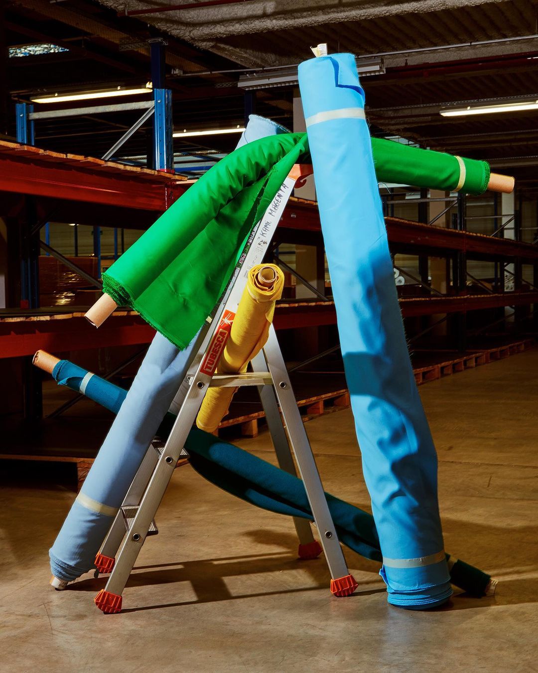 Assorted colorful fabric rolls are stacked on a silver step ladder inside a commercial storage facility with red and blue metal shelving.
