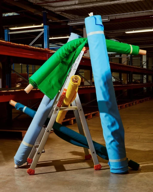 Assorted colorful fabric rolls are stacked on a silver step ladder inside a commercial storage facility with red and blue metal shelving.