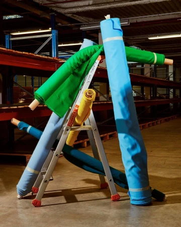 Assorted colorful fabric rolls are stacked on a silver step ladder inside a commercial storage facility with red and blue metal shelving.